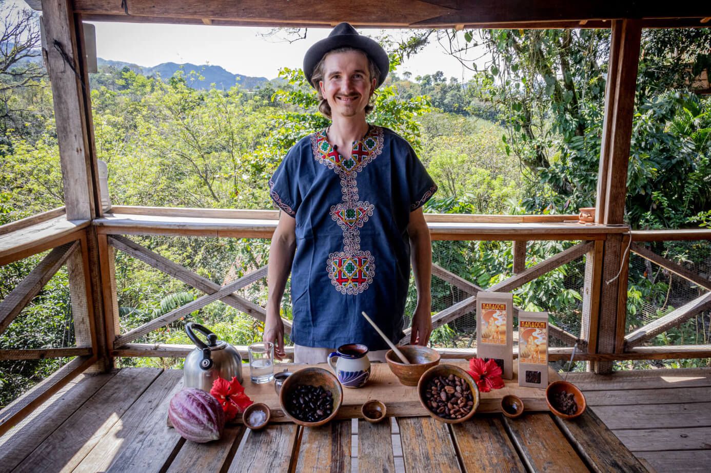 Mann mit Hut steht auf einer Holzveranda hinter einem Tisch mit Kakaobohnen, Tasse und Wasserkessel